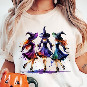 Könnte beinhalten: Beigefarbenes T-Shirt mit einer Aquarellillustration von drei Hexen. Jede Hexe trägt ein Kleid und einen spitzen Hut in Lila-, Blau- und Grüntönen. Das Design hat ein Halloween-Thema.