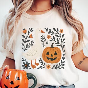 May include: Cream-colored t-shirt featuring a goose and jack-o'-lantern design. The goose is white with orange feet and beak, and the pumpkin is orange with a carved face. Black and orange floral accents surround the images, creating a Halloween theme.