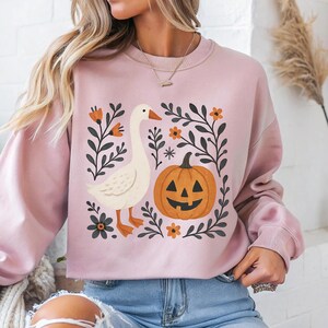 May include: A light pink sweatshirt featuring a whimsical Halloween design. The graphic includes a white goose, an orange jack-o'-lantern, and floral elements. The design is framed by dark green leaves and orange flowers, creating a festive autumn theme.