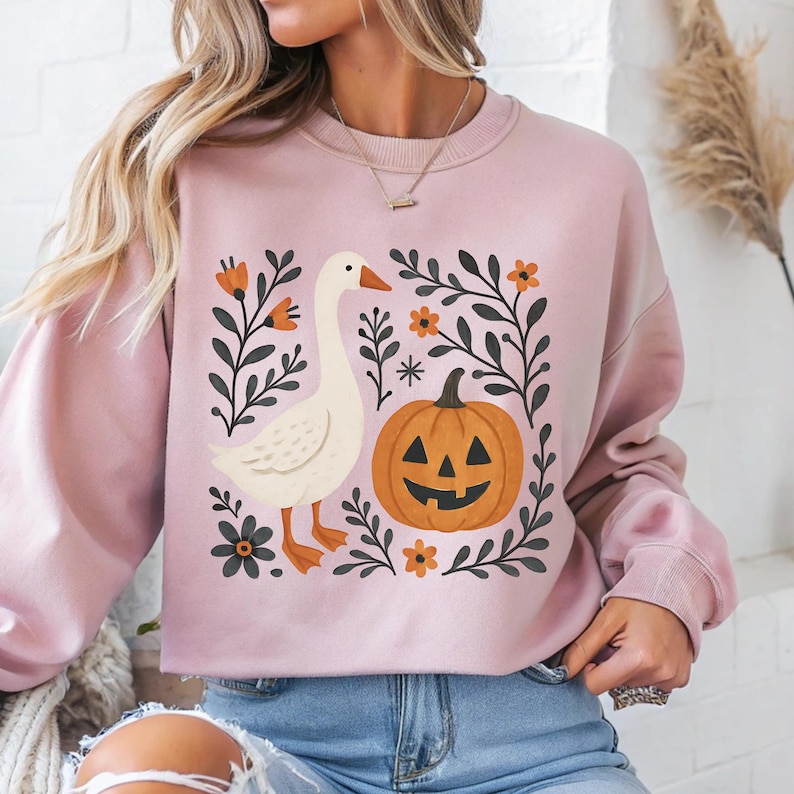 May include: A light pink sweatshirt featuring a whimsical Halloween design. The graphic includes a white goose, an orange jack-o'-lantern, and floral elements. The design is framed by dark green leaves and orange flowers, creating a festive autumn theme.