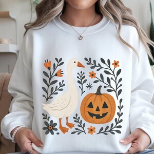 May include: White sweatshirt featuring a goose and a jack-o'-lantern design. The goose is cream-colored with orange feet and beak. The pumpkin is orange with a carved face. Black and orange floral elements surround the goose and pumpkin.