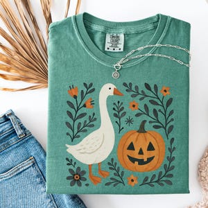 May include: A teal Comfort Colors t-shirt featuring a whimsical design. The graphic includes a white goose, an orange jack-o'-lantern, and floral elements. A silver necklace with a pendant rests on the shirt. The shirt is folded, with a pair of blue jeans visible.