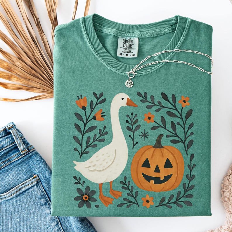 May include: A teal Comfort Colors t-shirt featuring a whimsical design. The graphic includes a white goose, an orange jack-o'-lantern, and floral elements. A silver necklace with a pendant rests on the shirt. The shirt is folded, with a pair of blue jeans visible.