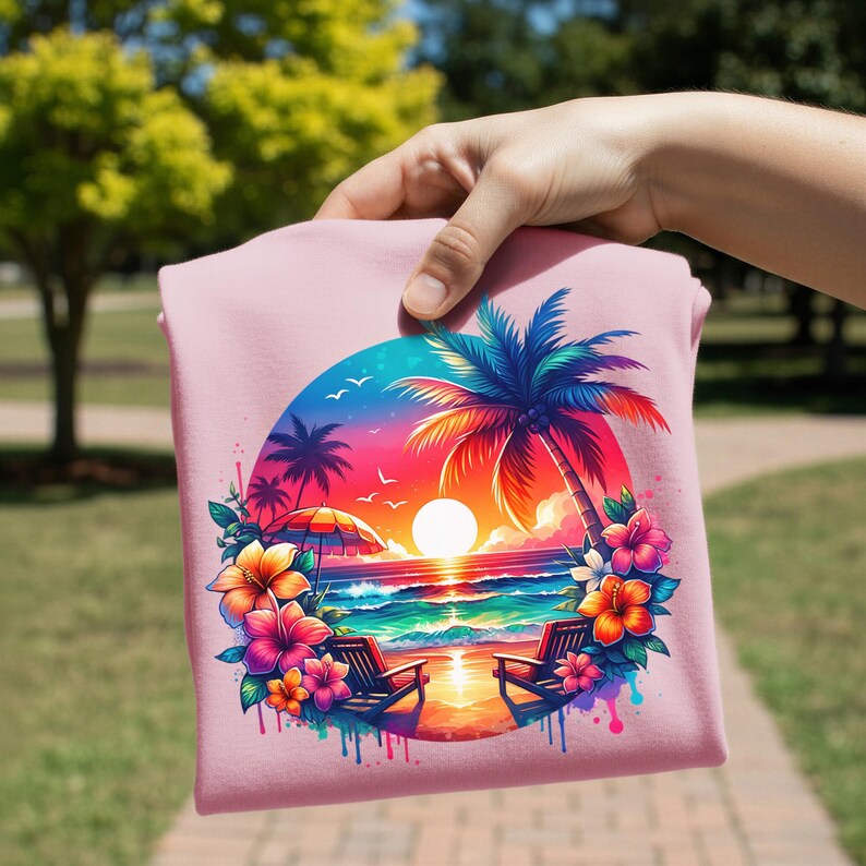 May include: A light pink t-shirt featuring a vibrant tropical sunset scene. The design includes palm trees, beach chairs, flowers, and a colorful sky. The image is held by a hand, showcasing the shirt's design.
