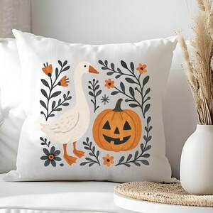 May include: White square throw pillow with a whimsical Halloween design. The pillow features a white goose, an orange jack-o'-lantern, and floral accents in orange and gray. The design is set against a white background.