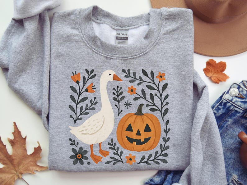 Transparent background PNG design featuring a white goose standing beside a smiling jack-o’-lantern pumpkin. The design includes black leafy botanical accents, orange flowers, a small star detail, and soft fall colors in a simple boho Halloween style.