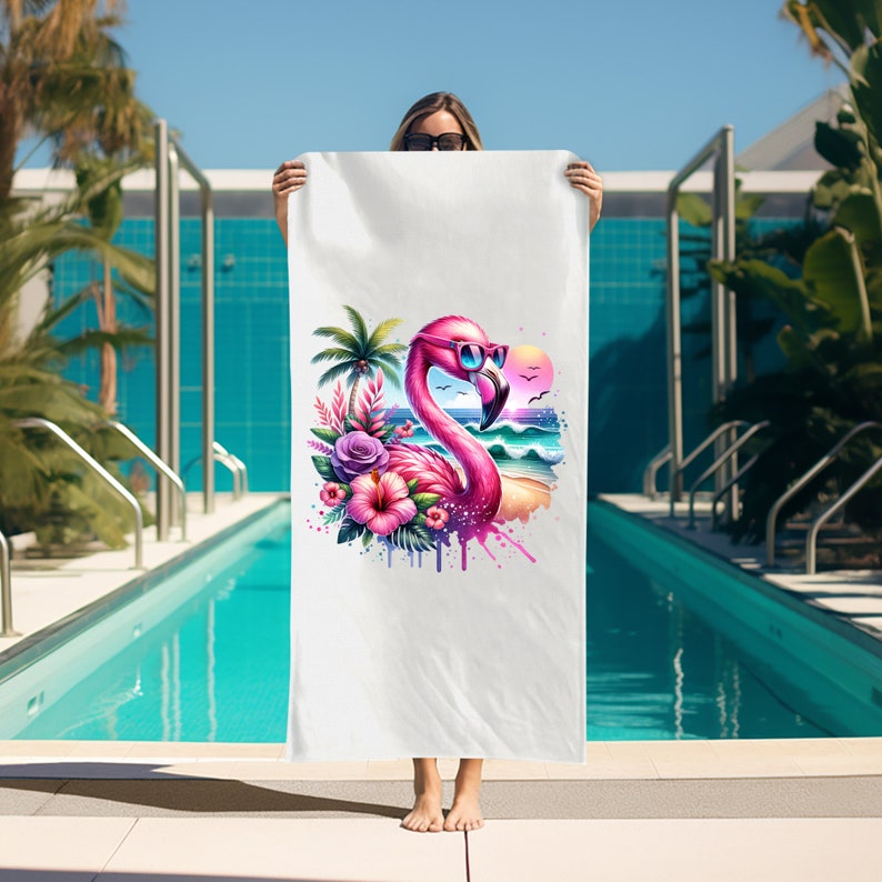 May include: A white beach towel with a pink flamingo wearing sunglasses and surrounded by tropical flowers and a palm tree. The flamingo is standing on a beach with waves and a sunset in the background.