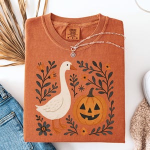 May include: Rust-colored Comfort Colors t-shirt with a graphic of a white goose, jack-o'-lantern, and floral elements. A silver necklace with a pendant is draped across the shirt. The shirt is folded on a white surface with denim jeans and a shoe visible.