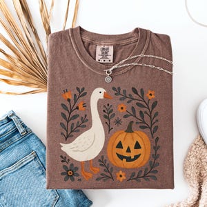 May include: Brown Comfort Colors t-shirt with a goose and jack-o'-lantern graphic. The design includes orange flowers and dark green foliage. A silver necklace with a sunburst pendant rests on the shirt. The shirt is folded next to blue jeans.