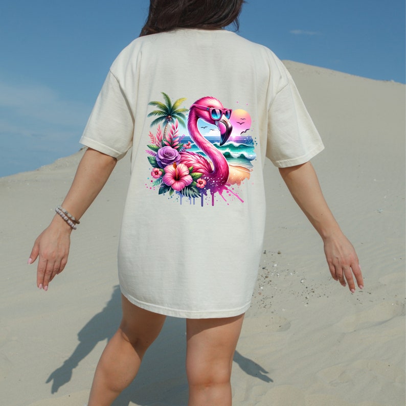 May include: A white t-shirt with a pink flamingo wearing sunglasses and surrounded by tropical flowers and a beach scene.