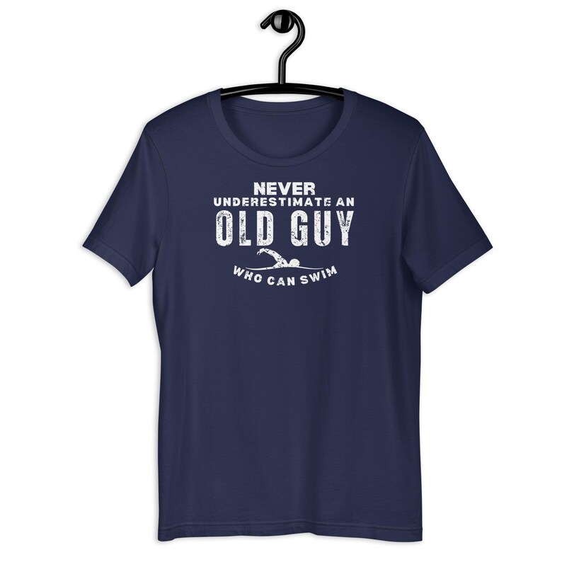 Puede incluir: Camiseta azul marino con el texto blanco "Never Underestimate an Old Guy Who Can Swim". El gr&aacute;fico incluye una ilustraci&oacute;n de nadador. La camiseta est&aacute; colgada en una percha negra.