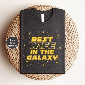 A black t-shirt with a yellow and white design that says "BEST WIFE IN THE GALAXY" and stars. The t-shirt is placed on a woven basket or plate.