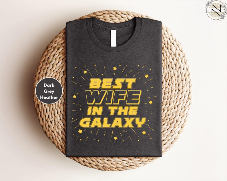 A black t-shirt with a yellow and white design that says "BEST WIFE IN THE GALAXY" and stars. The t-shirt is placed on a woven basket or plate.