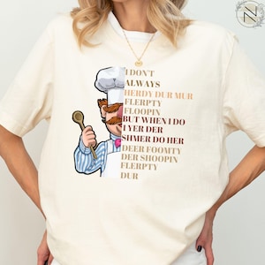 May include: Cream-colored t-shirt featuring a cartoon chef with a large mustache and chef's hat, holding a wooden spoon. The shirt has text that reads "I DON'T ALWAYS HERDY DUR MUR FLERPTY FLOOPIN BUT WHEN I DO I YER DER SHMER DO HER DEER FOOMTY DER SHOOPIN FLERPTY DUR."