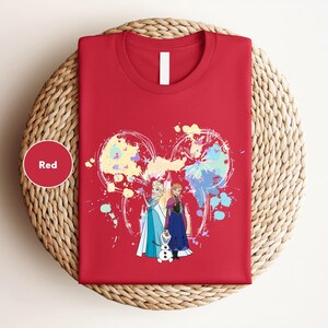 A red t-shirt with a colorful graphic design featuring two cartoon characters, one of which is a dog. The t-shirt is displayed on a woven basket or plate.