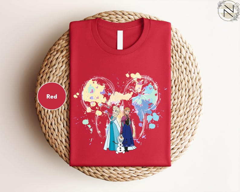 A red t-shirt with a colorful graphic design featuring two cartoon characters, one of which is a dog. The t-shirt is displayed on a woven basket or plate.