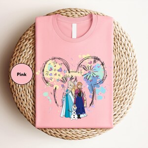 A pink t-shirt with a graphic design featuring two characters from the Disney movie Frozen, surrounded by colorful splashes and a Mickey Mouse-like design.