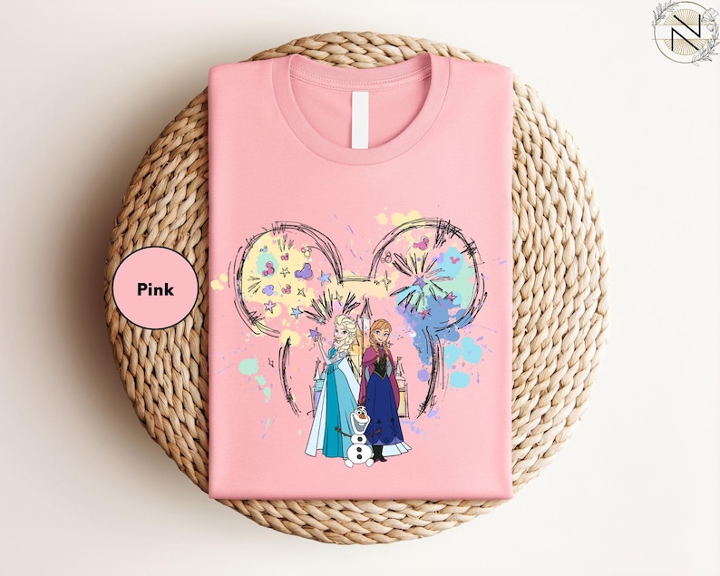 A pink t-shirt with a graphic design featuring two characters from the Disney movie Frozen, surrounded by colorful splashes and a Mickey Mouse-like design.