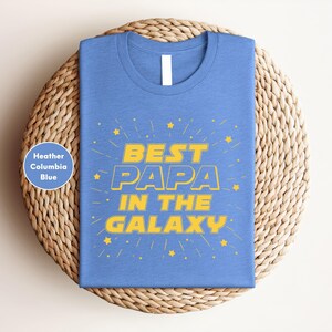 A blue t-shirt with the text "BEST PAPA IN THE GALAXY" printed on it, along with yellow stars. The t-shirt is displayed on a woven basket or plate.