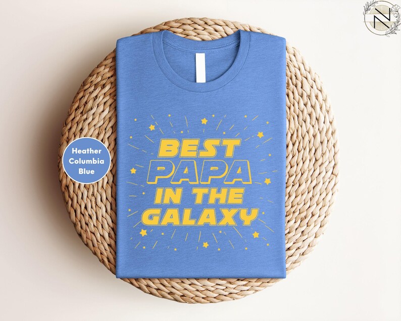 A blue t-shirt with the text "BEST PAPA IN THE GALAXY" printed on it, along with yellow stars. The t-shirt is displayed on a woven basket or plate.