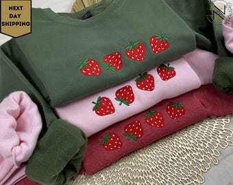 May include: A stack of three sweatshirts with embroidered strawberries. The sweatshirts are in shades of green, pink, and red.