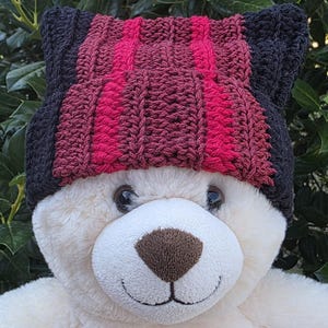 May include: A plush teddy bear wearing a handmade crochet hat. The hat features a striped pattern in shades of black, burgundy, and bright pink. The bear has a white body, a brown nose, and a smiling expression.