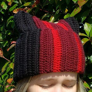 May include: A handmade crochet hat with cat ears. The hat features vertical stripes of black, red, and burgundy. The hat is made of a soft yarn and is designed to fit snugly on the head. The hat is a unique and stylish accessory.