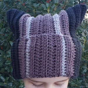 Puede incluir: Un gorro de ganchillo hecho a mano en tonos morado, gris y negro. El gorro presenta una textura acanalada y extensiones en forma de oreja en los laterales. El gorro está diseñado con rayas verticales de varios colores.