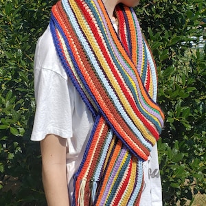 May include: A colorful striped crocheted scarf with fringe. The scarf is made with a variety of colors, including red, orange, yellow, green, blue, and purple.