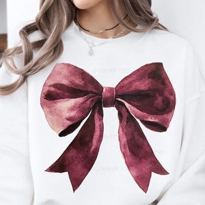 May include: White sweatshirt featuring a large watercolor bow in shades of burgundy and pink. The bow is centered on the front of the shirt. The text "ENCHANTED ECHO" is visible on the left side of the bow.