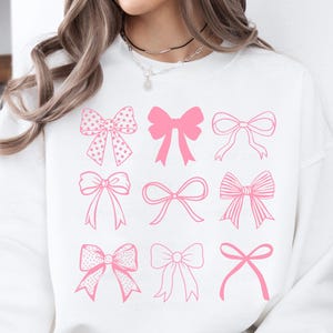 May include: White sweatshirt featuring a pink bow design. The design includes nine different bow illustrations in various styles, including polka dots and solid outlines. The bows are arranged in a grid pattern across the front of the sweatshirt.