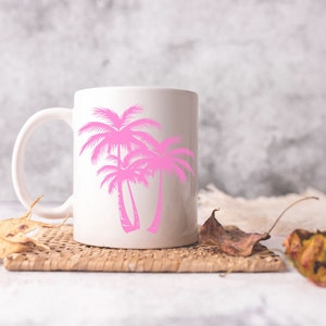 K&ouml;nnte beinhalten: Eine wei&szlig;e Keramiktasse mit einem rosafarbenen Palmenbaum-Design. Die Tasse steht auf einem geflochtenen Untersetzer mit getrockneten Bl&auml;ttern.