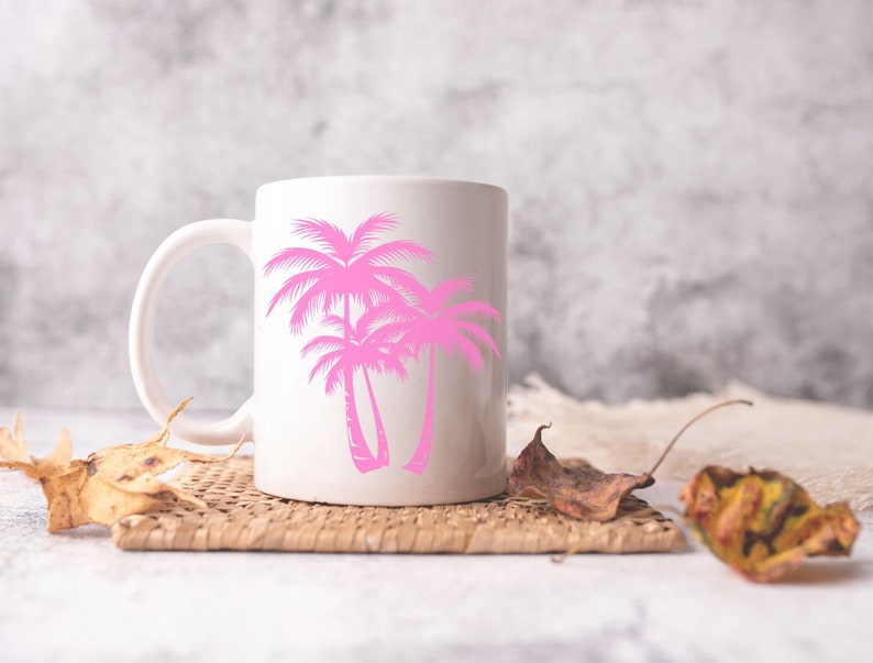 K&ouml;nnte beinhalten: Eine wei&szlig;e Keramiktasse mit einem rosafarbenen Palmenbaum-Design. Die Tasse steht auf einem geflochtenen Untersetzer mit getrockneten Bl&auml;ttern.