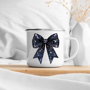 May include: White enamel mug with a black glitter bow design. The mug has a handle and a white background.