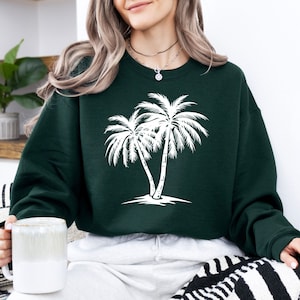 K&ouml;nnte beinhalten: Ein dunkelgr&uuml;ner Pullover mit einem wei&szlig;en Palmenbaum-Motiv. Der Pullover hat einen Rundhalsausschnitt und lange &Auml;rmel.