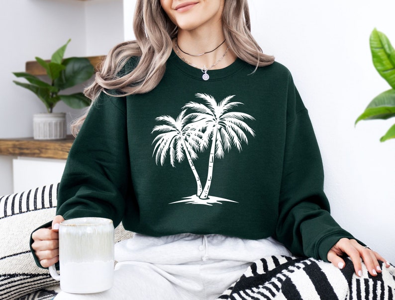 K&ouml;nnte beinhalten: Ein dunkelgr&uuml;ner Pullover mit einem wei&szlig;en Palmenbaum-Motiv. Der Pullover hat einen Rundhalsausschnitt und lange &Auml;rmel.
