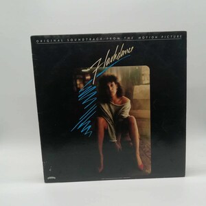 May include: A vinyl record album cover for the movie "Flashdance." The cover is black with the movie title in a stylized, turquoise font. A woman is seated in a dimly lit space, wearing a gray top and red shoes.