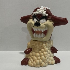 Puede incluir: Figura del Demonio de Tasmania con una sonrisa amenazante, grandes dientes blancos y una nariz negra. La figura tiene orejas marrones y un cuerpo beige con una base texturizada de color marrón claro. Objeto de colección.