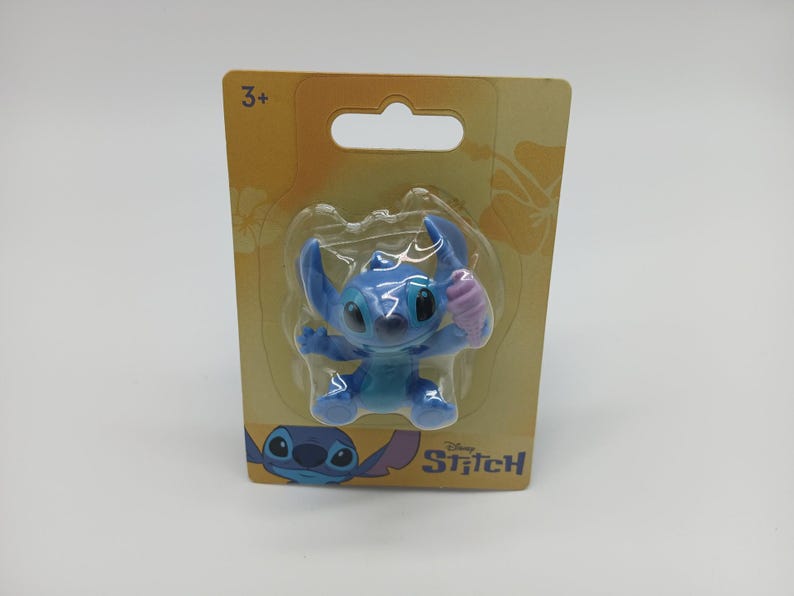 May include: A blue Stitch figurine holding a pink ice cream cone, packaged on a yellow card with the Disney Stitch logo. The packaging includes a "3+" age recommendation.