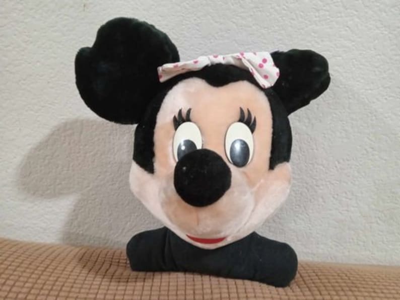 May include: A plush toy of Mickey Mouse's girlfriend Minnie Mouse. She is wearing a pink and white polka dot bow and has black ears and a black nose. Her eyes are closed and she has a big smile.