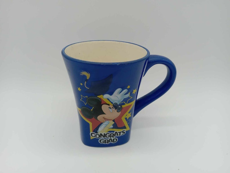 May include: Blue ceramic mug with a white interior and a curved handle. The mug features Mickey Mouse in a graduation cap and gown, with the words "Congrats Grad" and star accents. A celebratory gift.