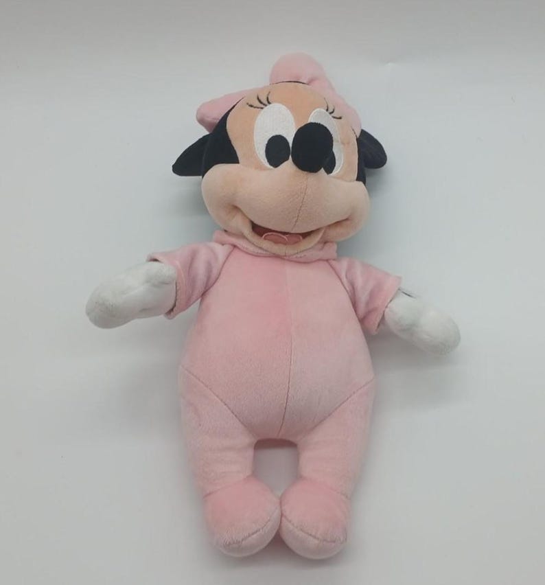 Peut inclure: Une poup&eacute;e en peluche Minnie Mouse v&ecirc;tue d'une barboteuse rose clair et d'un n&oelig;ud assorti. La poup&eacute;e a des oreilles noires, un visage rose et des gants blancs. Un jouet doux et c&acirc;lin.