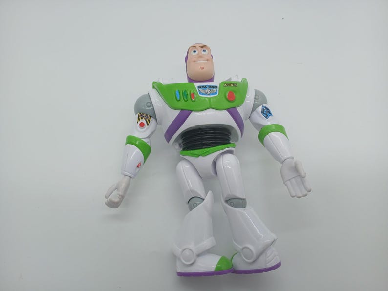 May include: A Buzz Lightyear action figure, featuring a white and green spacesuit with purple accents. The toy has a smiling face, articulated limbs, and a green chest panel with buttons. The figure is standing on a white surface.