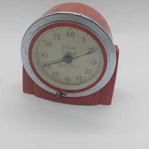 May include: A vintage red Kodak Timer with a silver bezel. The timer has a white face with black numerals and hands, and the words "KODAK TIMER" are printed on the face. The timer is approximately 4 inches tall.
