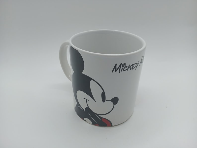 May include: White ceramic mug with a black and red Mickey Mouse design. The mug has a curved handle and the words "Mickey Mouse" in black script. A classic Disney mug for hot beverages.