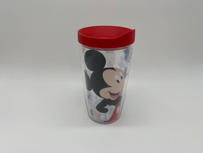May include: Clear plastic tumbler with a red lid featuring a smiling Mickey Mouse graphic. The tumbler has a double-walled design and is decorated with paw prints. A fun and functional drinkware item.