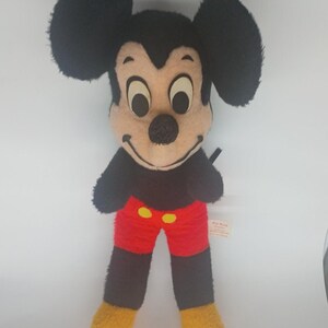 May include: Vintage Mickey Mouse plush toy. The plush features a black head and ears, a tan face, black arms, red shorts with yellow buttons, and yellow feet. The toy is standing upright against a white background.