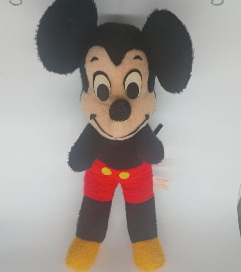 May include: Vintage Mickey Mouse plush toy. The plush features a black head and ears, a tan face, black arms, red shorts with yellow buttons, and yellow feet. The toy is standing upright against a white background.