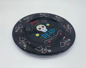 May include: Black ceramic plate with a Day of the Dead theme. It features a sugar skull with colorful flowers and the text "Day of the Dead" with dancing skeletons around the border.