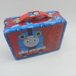 May include: A rectangular, red and blue metal lunchbox featuring the cartoon character Thomas the Tank Engine. The lunchbox has a red handle and the words "ALL ABOARD" are printed on the side.